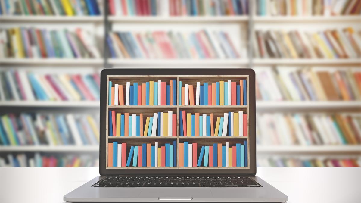 A laptop sitting on a table showing colourful library books on its screen, against a backdrop of books on library shelves.