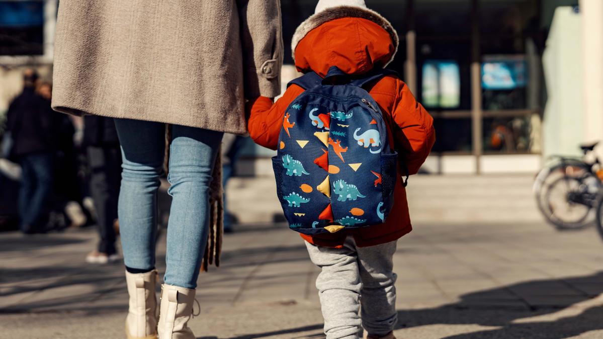 A rear view of a woman wearing a tan car coat walking toward the entrance of a schoolwith her kindergarten aged child who is wearing a backpack covered in animal cartoons.