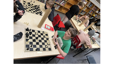 Children playing chess in a library at multiple tables.