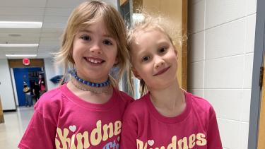 Two primary grade girls wearing pink shirts holding hands and smiling in a school hallway.