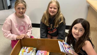BKES Book Swap! Three girls seated around a cardboard box of books
