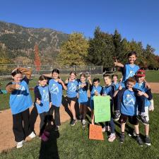 Elementary students in light and dark blue shirts stand together in a green field on a sunny fall day following a cross-country running event